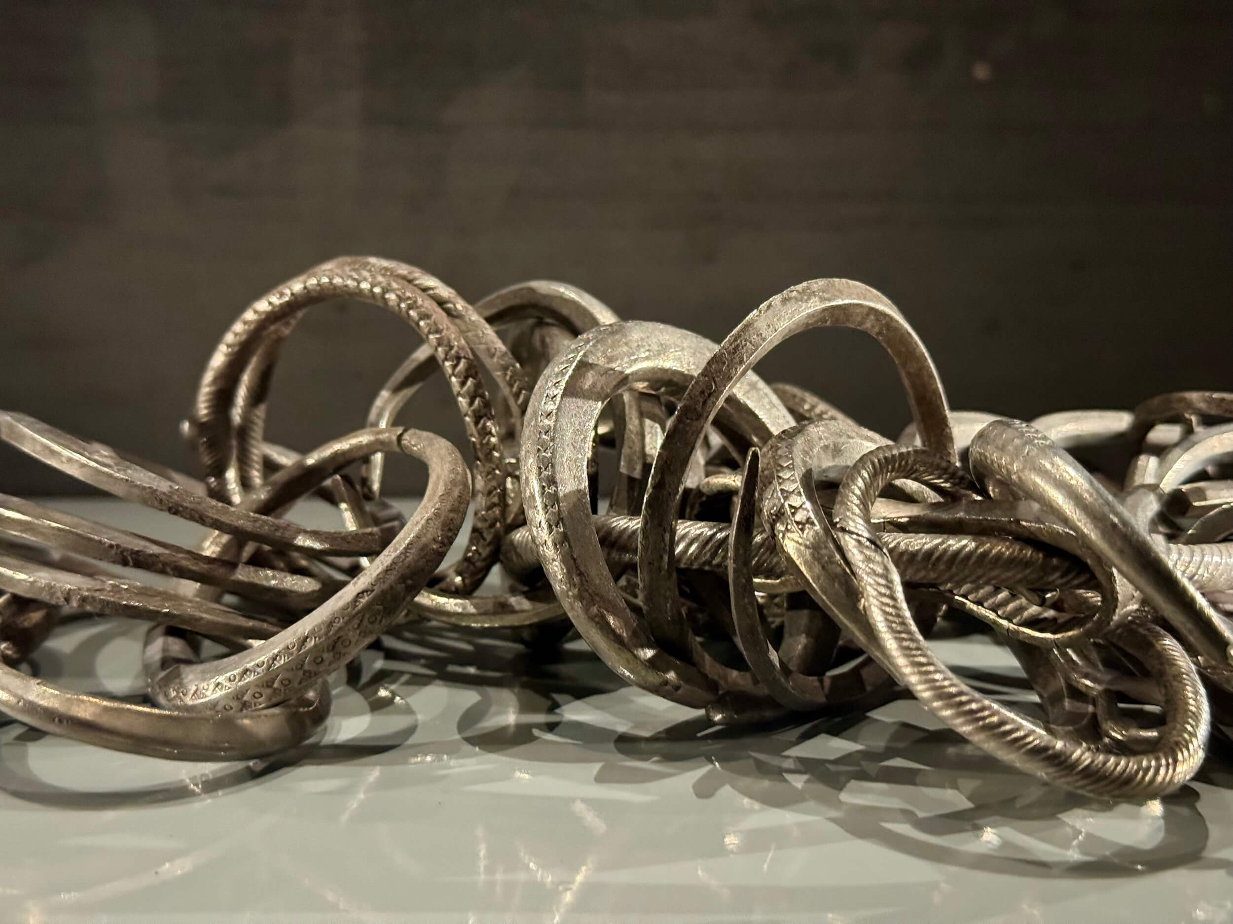 En närbild av flera sammanflätade och överlappande silverarmband eller armringar, med graverade och texturerade mönster, visade på en reflekterande yta mot en mörk bakgrund.