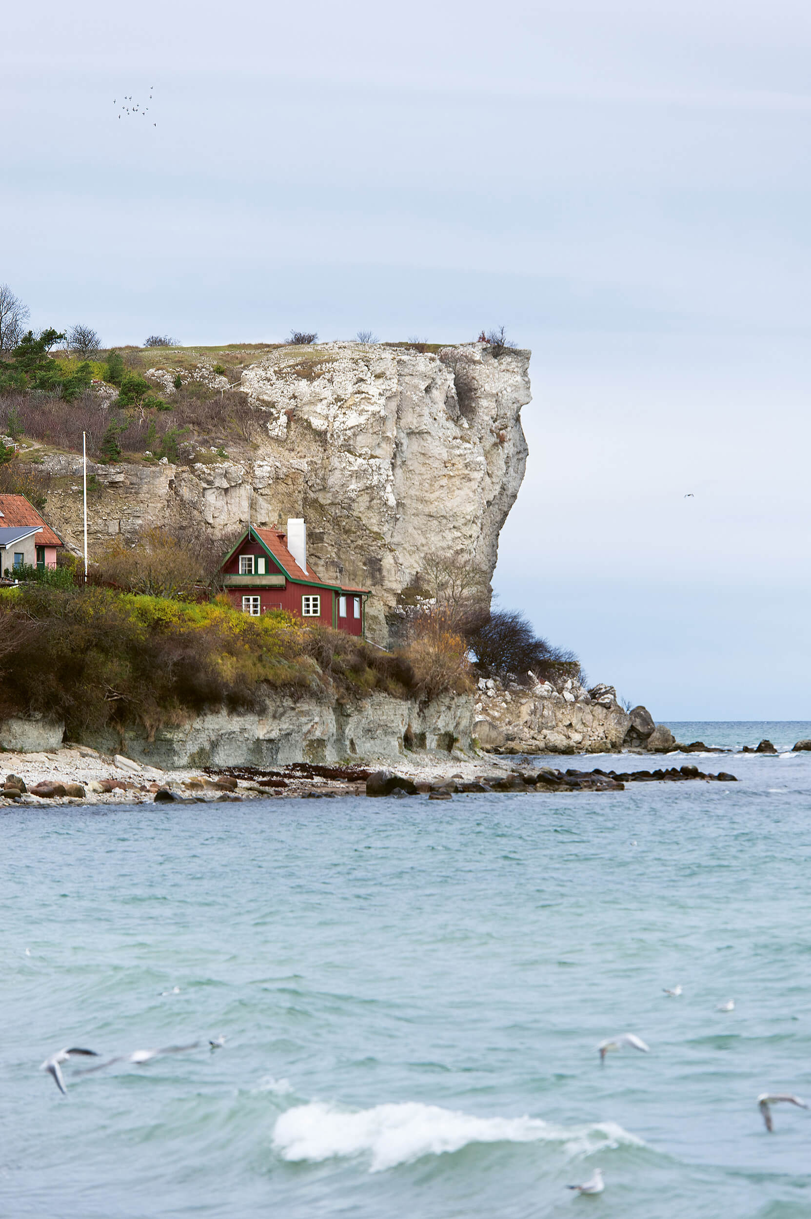 Ett litet rött hus med brant tak ligger på en gräsbevuxen klippa vid havet, med en stor vit klippvägg som reser sig bakom och vågor i förgrunden. Måsar flyger över vattnet under en molnig himmel.