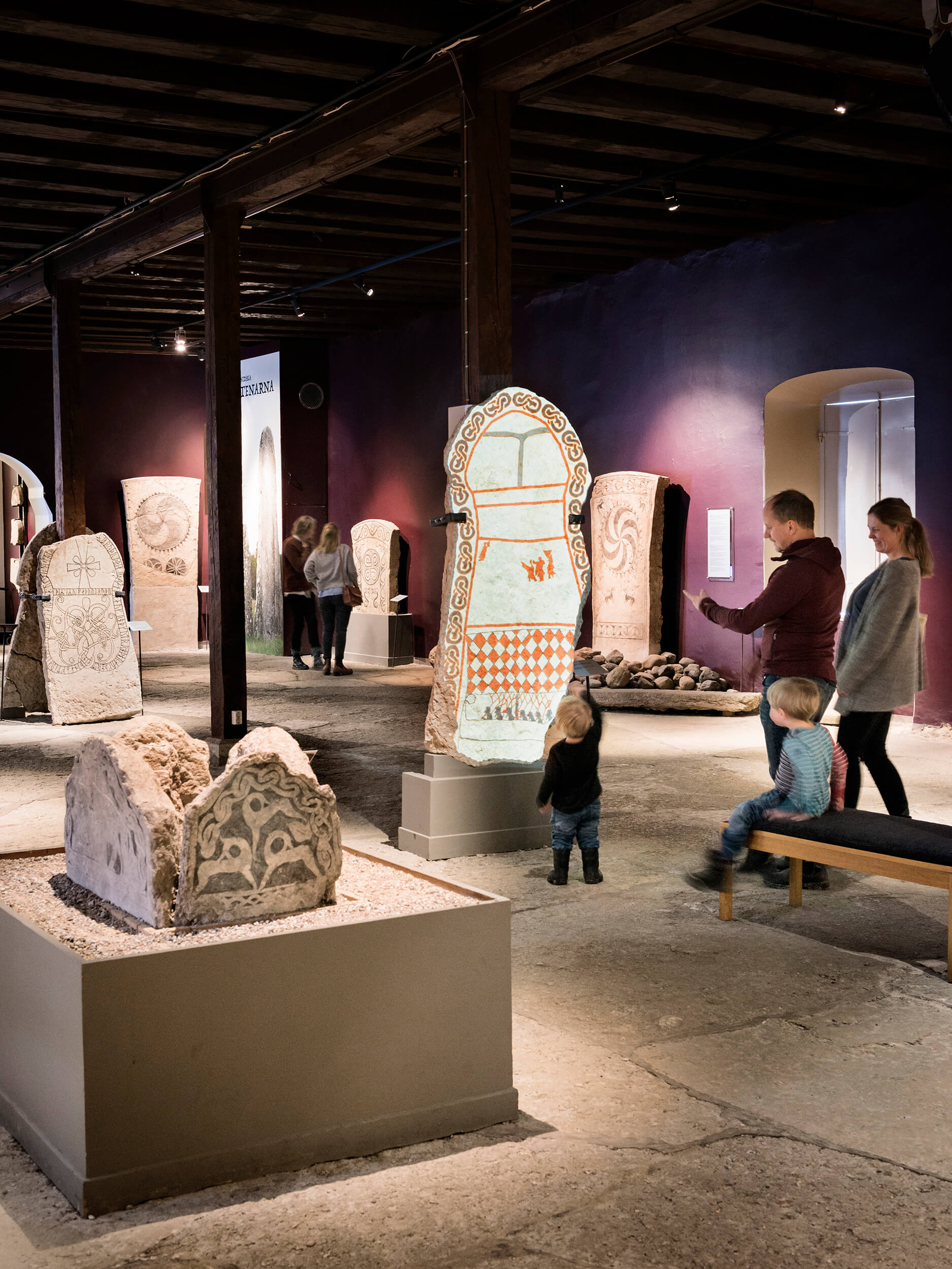 Besökare, inklusive en familj med två små barn, utforskar ett museumsgalleri som visar antika snidade stenartefakter och runstenar på piedestaler under mjuk belysning.