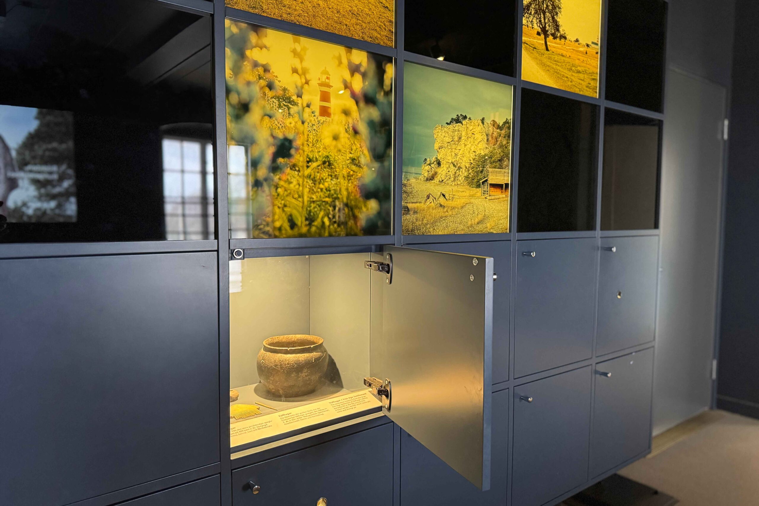 En museimonter med fyrkantiga fack; ett öppet fack avslöjar ett gammalt keramikkärl, medan andra har landskapsfoton och stängda dörrar. Ljus framhäver keramiken mot en mörk rumsbakgrund.