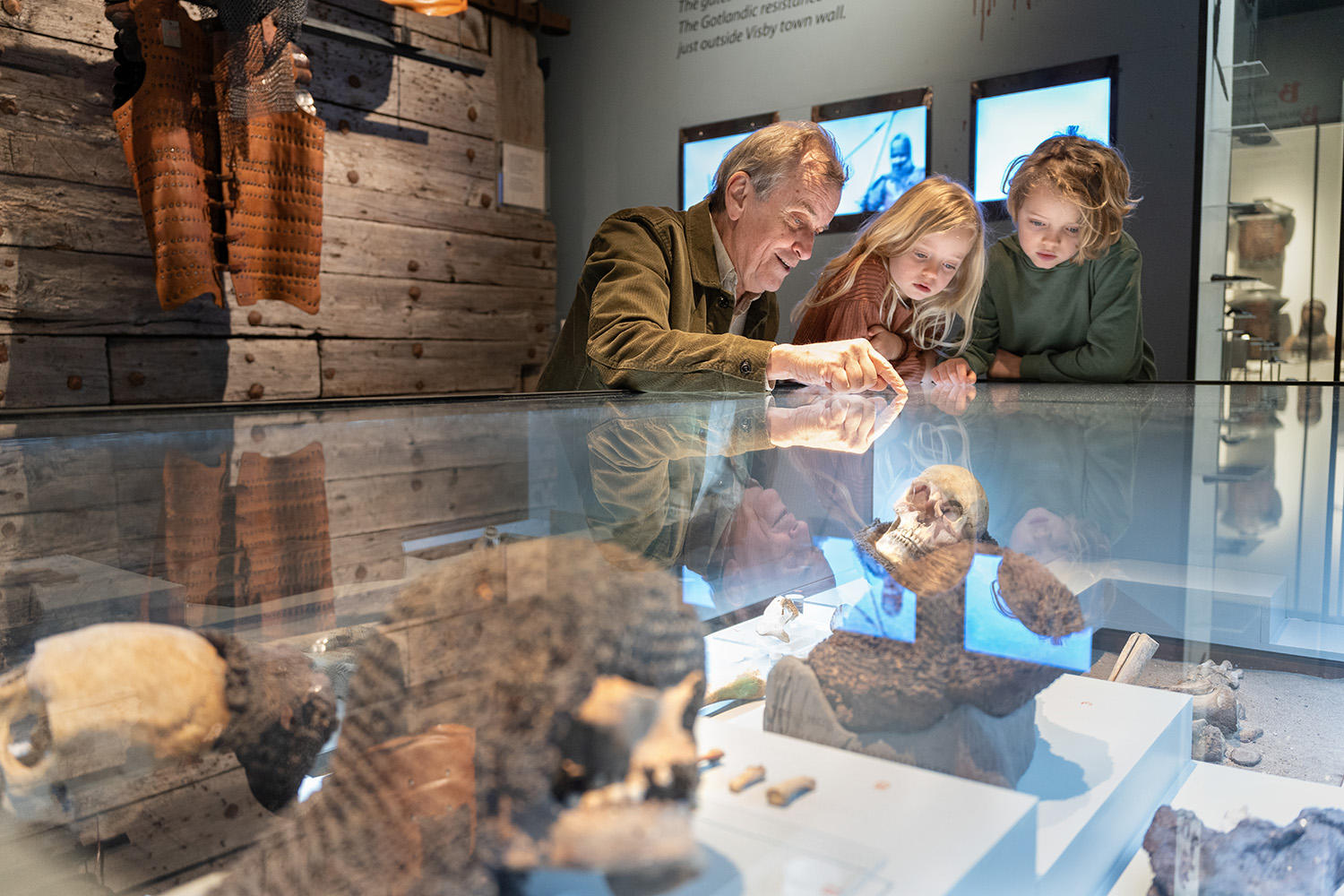En äldre man och två barn tittar noga på artefakter som visas i en glasmonter på ett museum. Reflektioner av föremålen och människorna syns på glasytan.