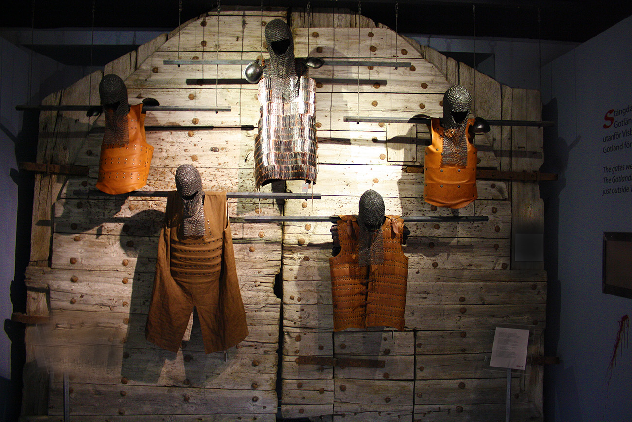 Utställning med fem historiska rustningar med ringbrynjshjälmar och skyddsvästar som hänger på en stor trävägg i en museiutställning, med informationsplaketter synliga på sidorna.