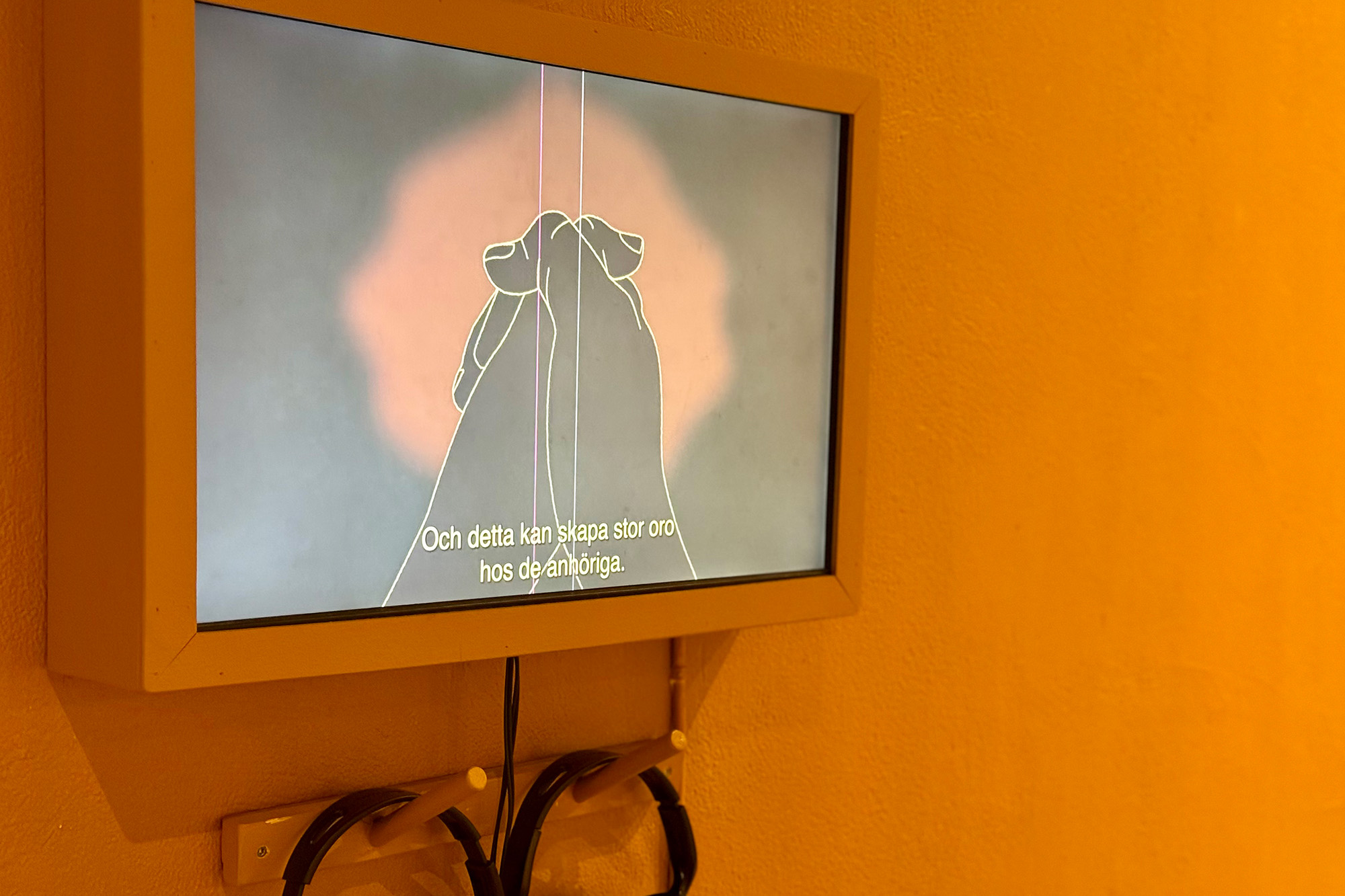 En väggmonterad skärm visar en animation av två kor med svenska undertexter. Hörlurar hänger under skärmen och väggen är målad gul.