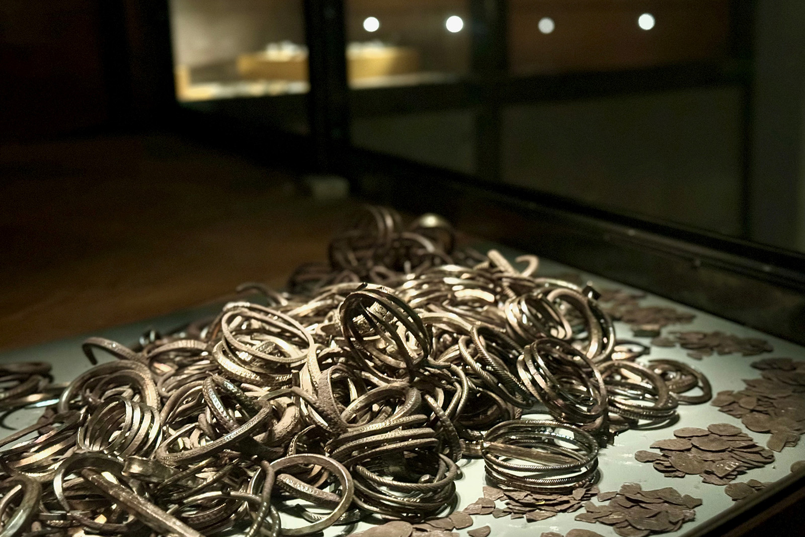 En hög med metallarmband och spridda mynt utställda i en glasmuseimonter, med en svagt upplyst bakgrund som visar upp fler artefakter i suddiga glashöljen.