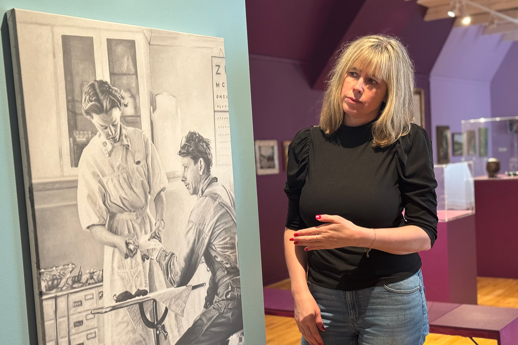 En kvinna med blont hår står bredvid en svartvit teckning av en medicinsk scen i ett museum eller galleri med lila väggar och tittar fundersamt på konstverket.