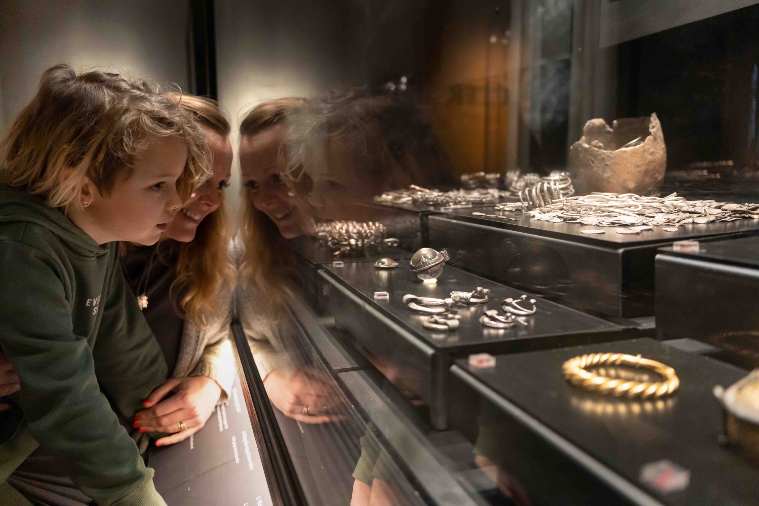 Ett barn och en vuxen undersöker noggrant antika smycken och artefakter som visas i en glasmonter på ett museum, deras reflektioner syns i glaset.