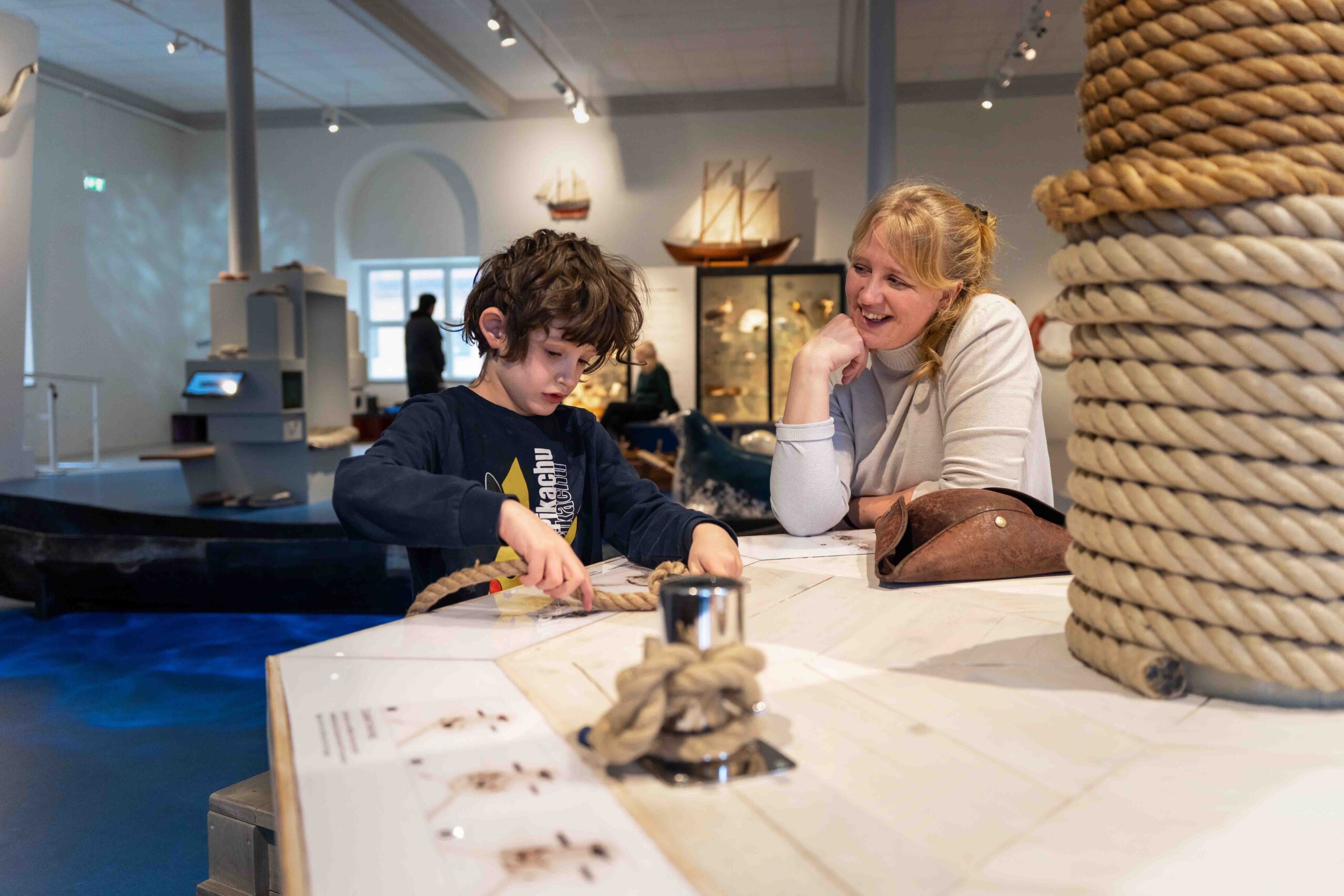 En ung pojke övar på att knyta nautiska knopar på en museiutställning medan en kvinna, troligen hans mamma, tittar på och ler. Nautiska rep och maritima utställningar syns i bakgrunden.