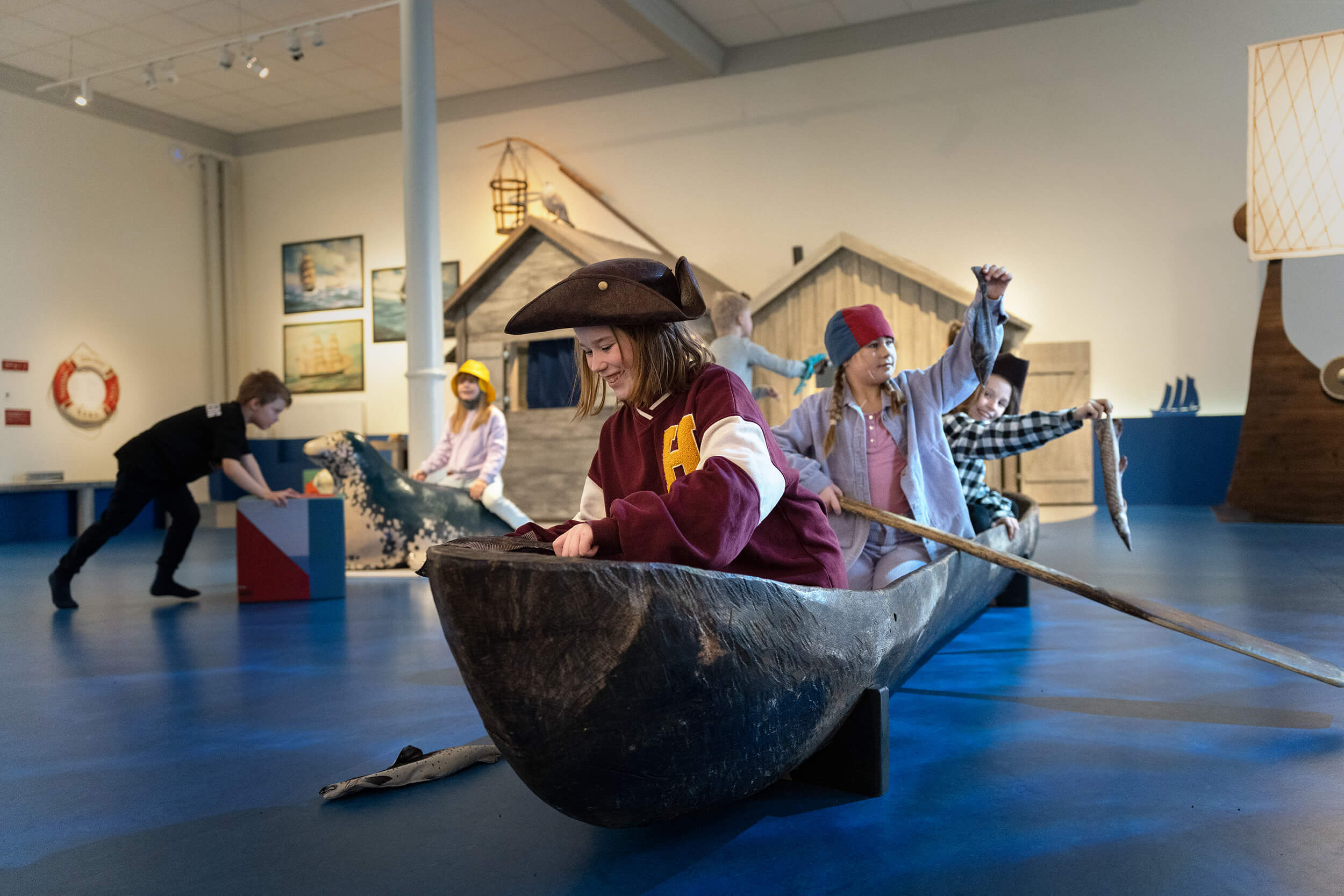Barn leker i en museiutställning och låtsas ro en träbåt i pirat- och sjömanskostymer. I bakgrunden interagerar andra barn med interaktiva utställningar och rekvisitahus, vilket skapar en lekfull och fantasifull scen.