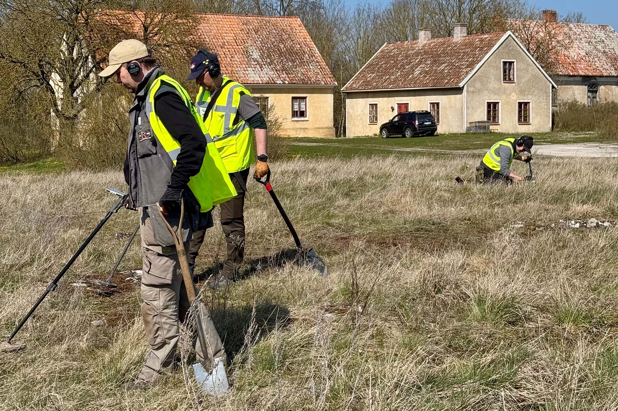 Tre personer som går med metallsökare. En gård i bakgrunden.