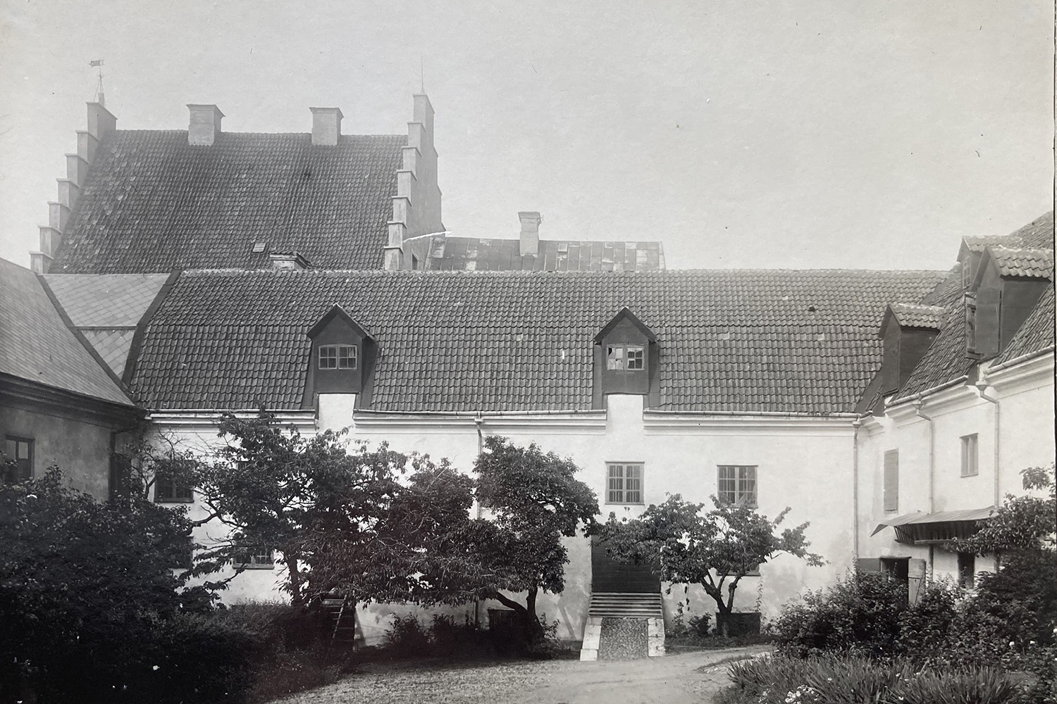Svartvitt foto av en historisk byggnad med tegeltak, takkupor och en central trappa, omgiven av träd och en innergård. Ett större tak med skorstenar syns i bakgrunden.