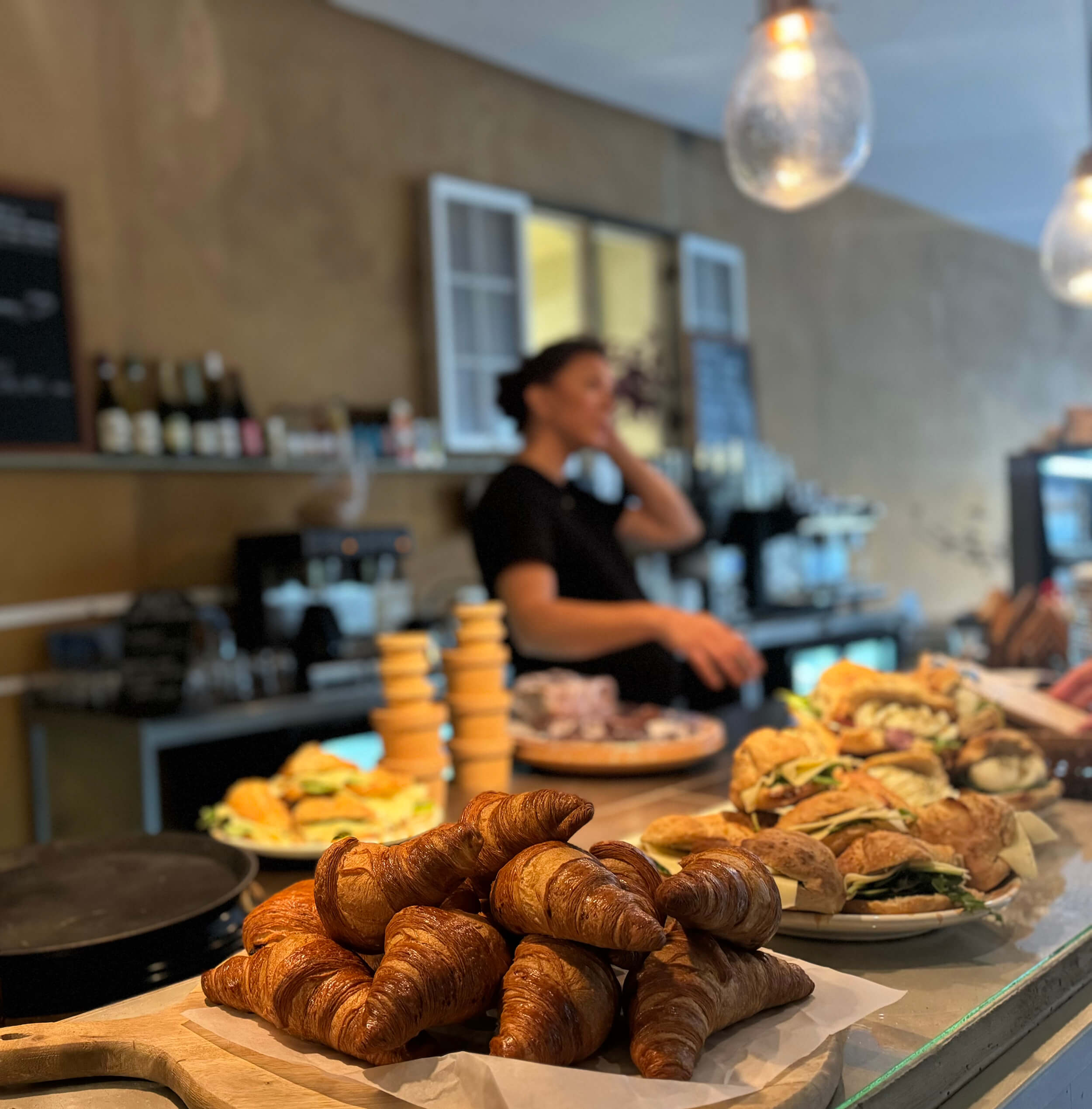 En tallrik croissanter står på en disk i ett mysigt kafé, med smörgåsar och bakverk i närheten. I bakgrunden pratar en person i telefon nära hyllor med flaskor och tallrikar. Scenen är varmt upplyst.