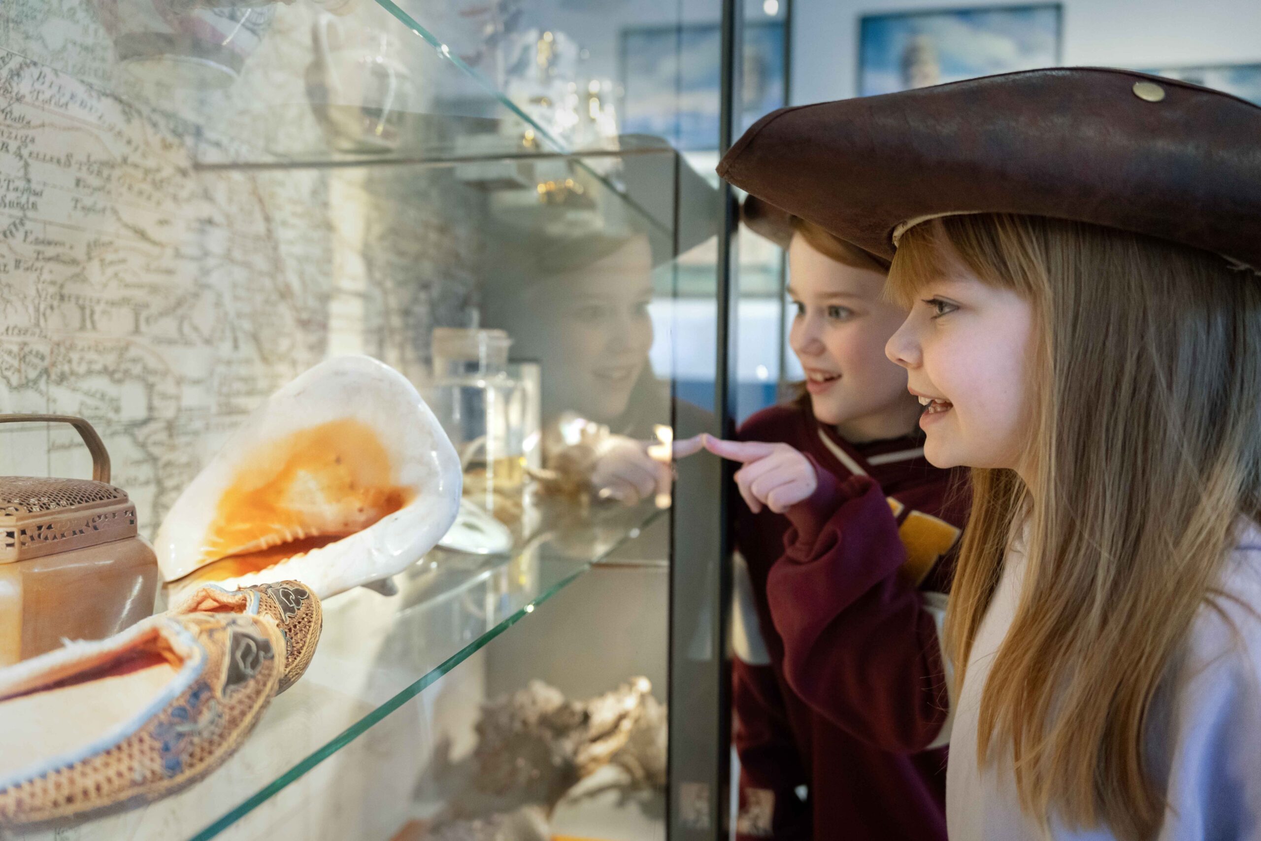 Två unga flickor, en med pirathatt, ler och pekar entusiastiskt på snäckskal och maritima föremål som visas bakom glas i en museiutställning.