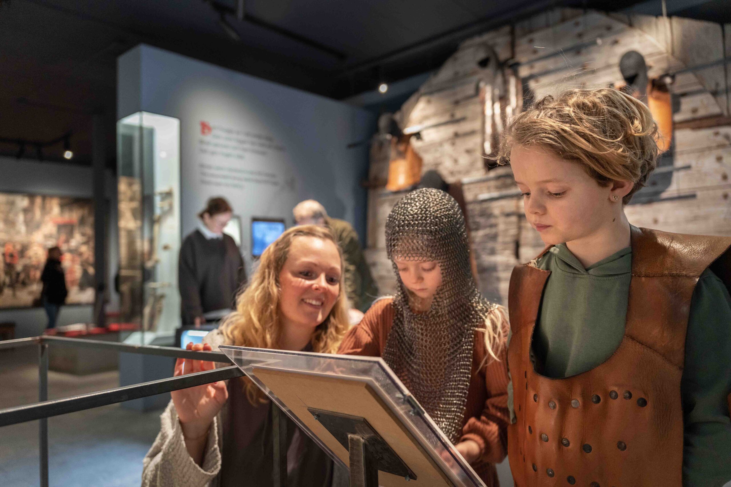En kvinna och två barn, ett i ringbrynja och det andra i brun väst, engagerar sig i en interaktiv utställning på ett museum med medeltida artefakter i bakgrunden.