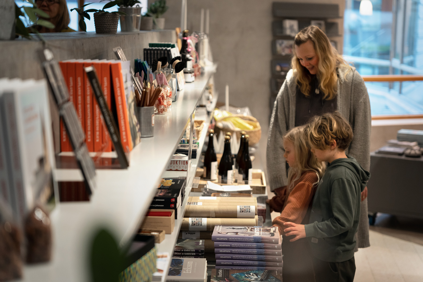 En kvinna och två barn står tillsammans i en mysig butik och tittar på böcker och kontorsmaterial på en väl upplyst hylla. Barnen verkar intresserade av varorna, medan kvinnan ler och tittar på dem.