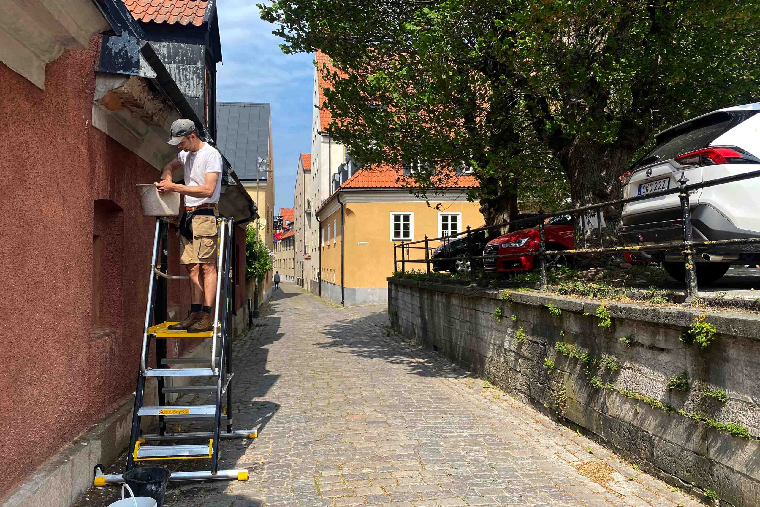 En man står på en stege och målar den övre delen av en texturerad röd vägg längs en kullerstensgränd. Parkerade bilar och träd kantar den upphöjda trottoaren till höger, med gula byggnader synliga i bakgrunden.