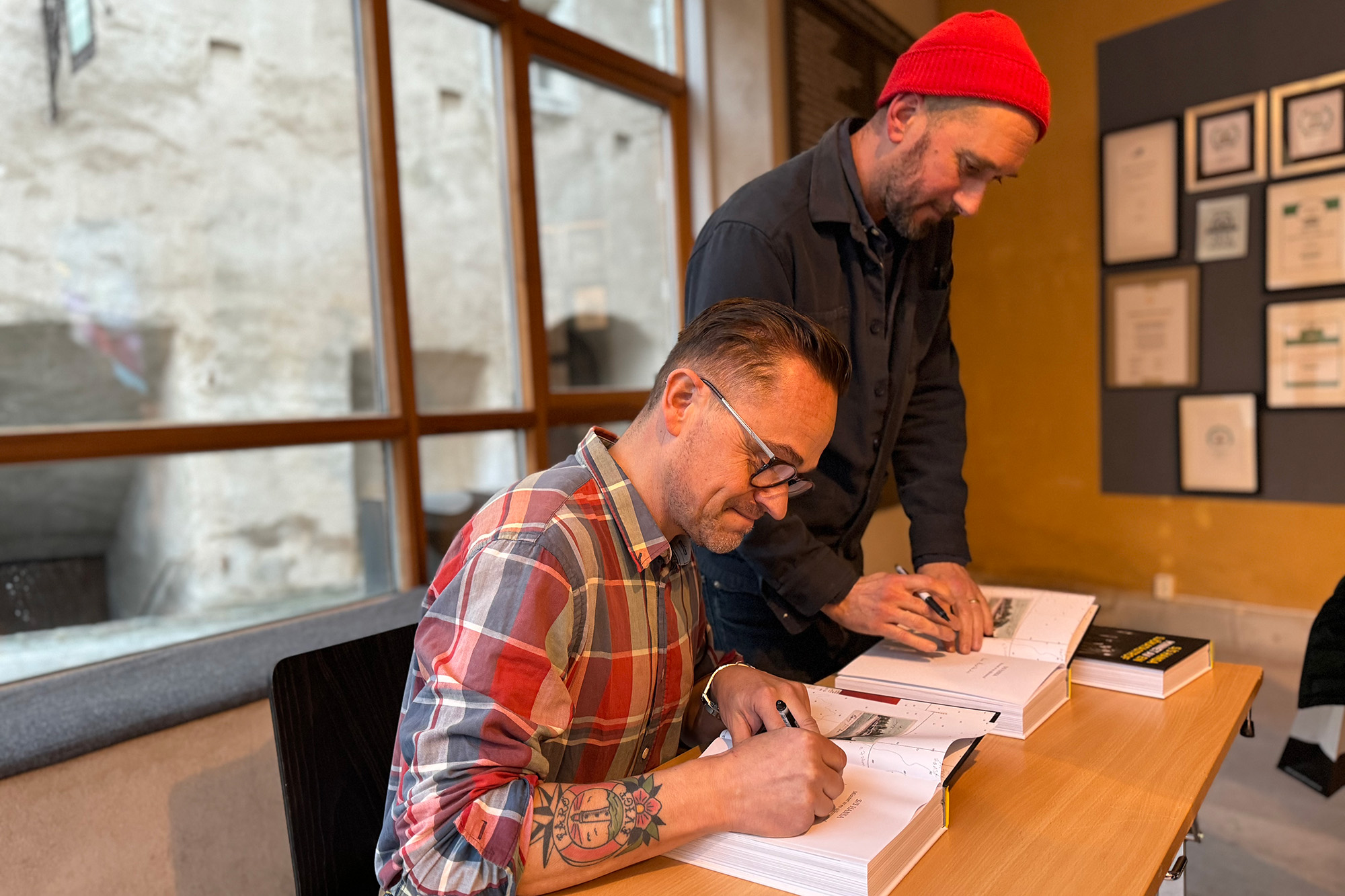 Två män signerar böcker vid ett bord inomhus. Mannen framför bär glasögon och rutig skjorta, medan mannen bakom honom bär röd mössa och mörk jacka. Inramade bilder och certifikat hänger på den gula väggen.