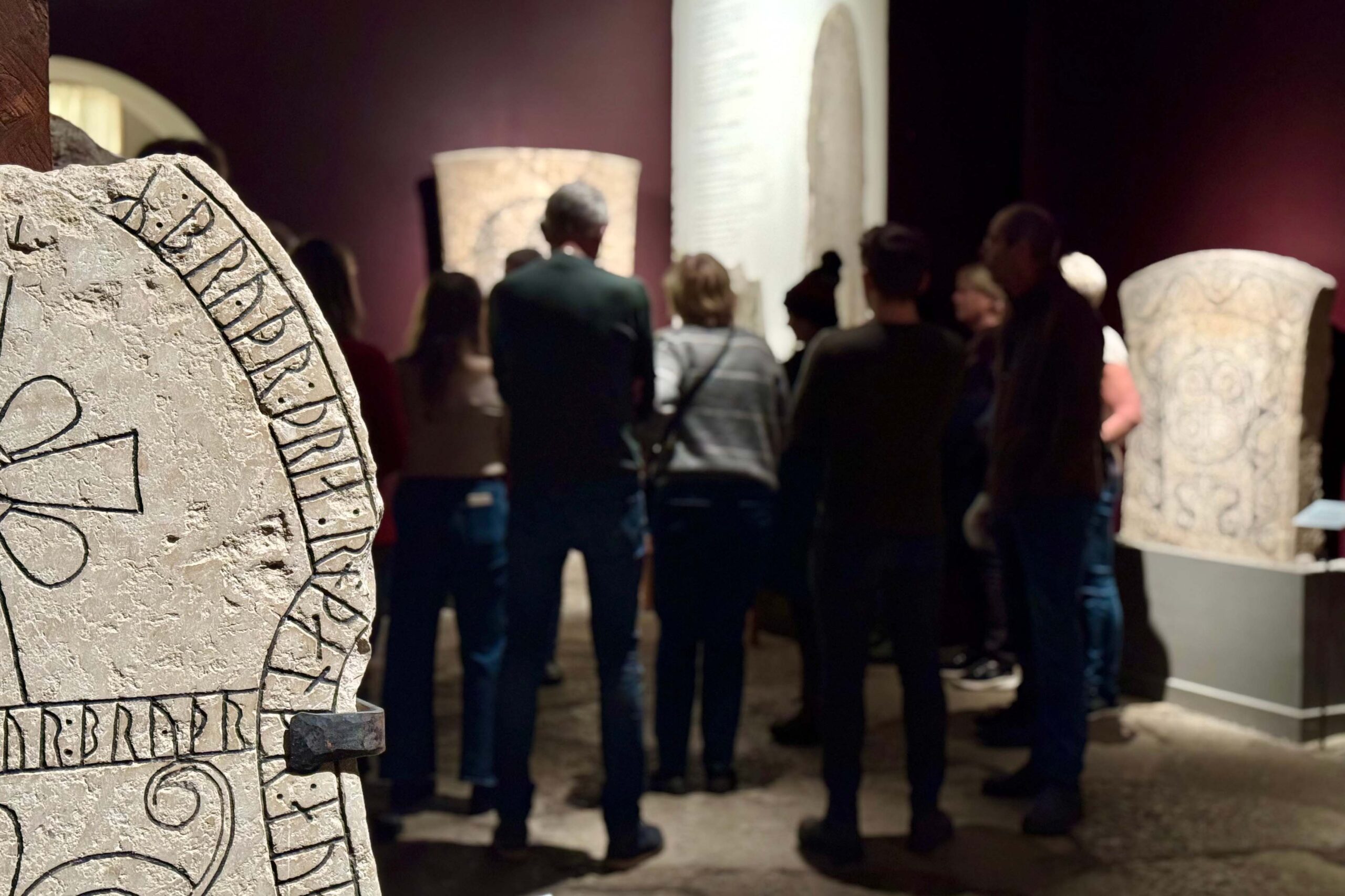 En grupp människor står i ett svagt upplyst museirum och lyssnar på en guide. I förgrunden syns en runsten med inristade nordiska runor tydligt.