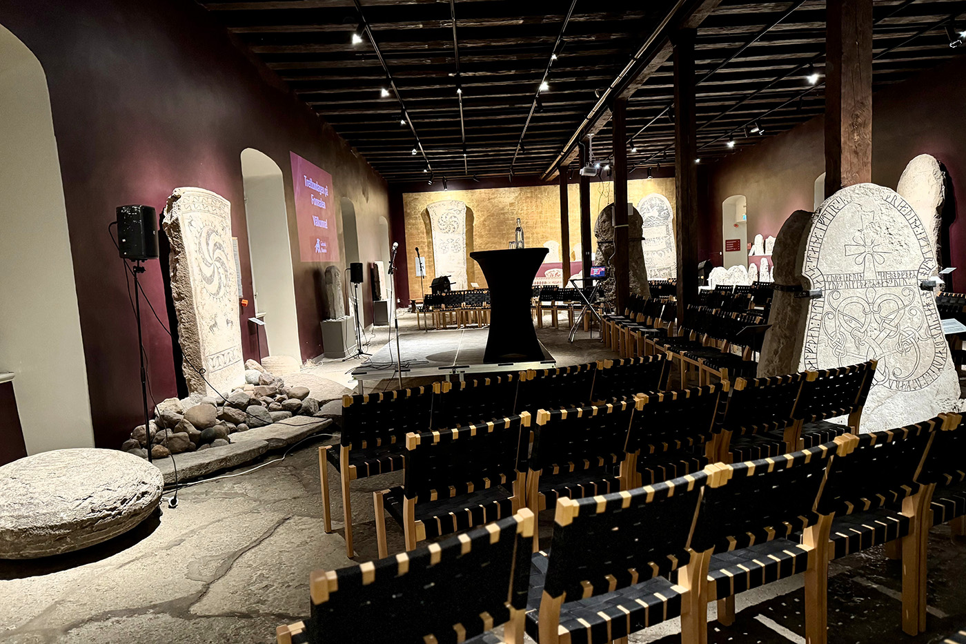 Rader av svarta stolar vetter mot en scen i ett svagt upplyst rum med antika runstenar och artefakter utställda mot rödbruna och guldfärgade väggar, vilket skapar en museiliknande eller historisk evenemangsmiljö.