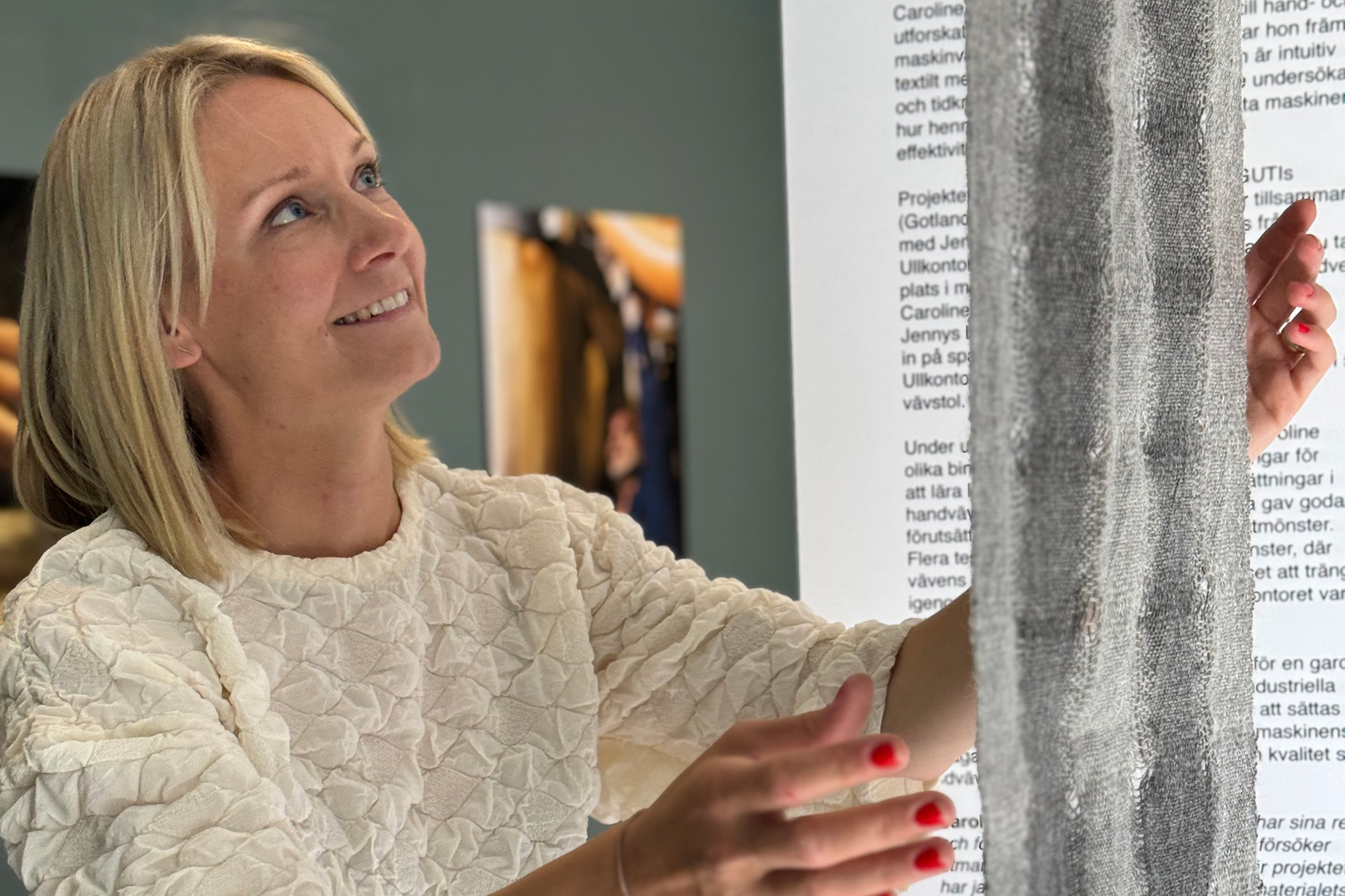 En kvinna med blont hår och en vit texturerad topp ler medan hon undersöker eller rör vid ett vertikalt stycke grått tyg. Text tryckt på en närliggande vägg syns i bakgrunden.