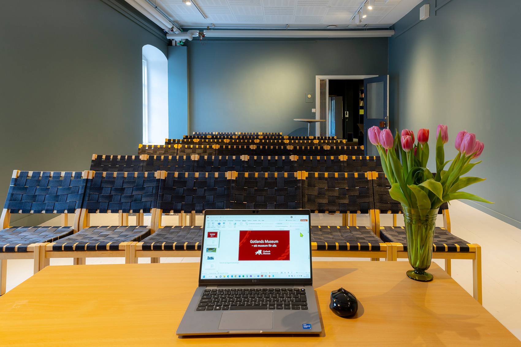 En bärbar dator och en vas med rosa tulpaner står på ett träskrivbord framför tomma rader av flätade stolar i ett väl upplyst, modernt konferensrum.