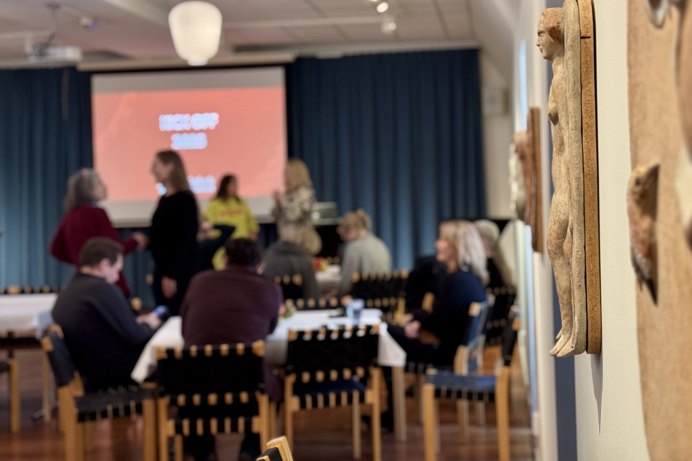 En grupp människor samlas i ett rum med bord och stolar, en presentationsskärm i bakgrunden och en träskulptur av en person i förgrunden. Atmosfären verkar vara som ett möte eller seminarium.