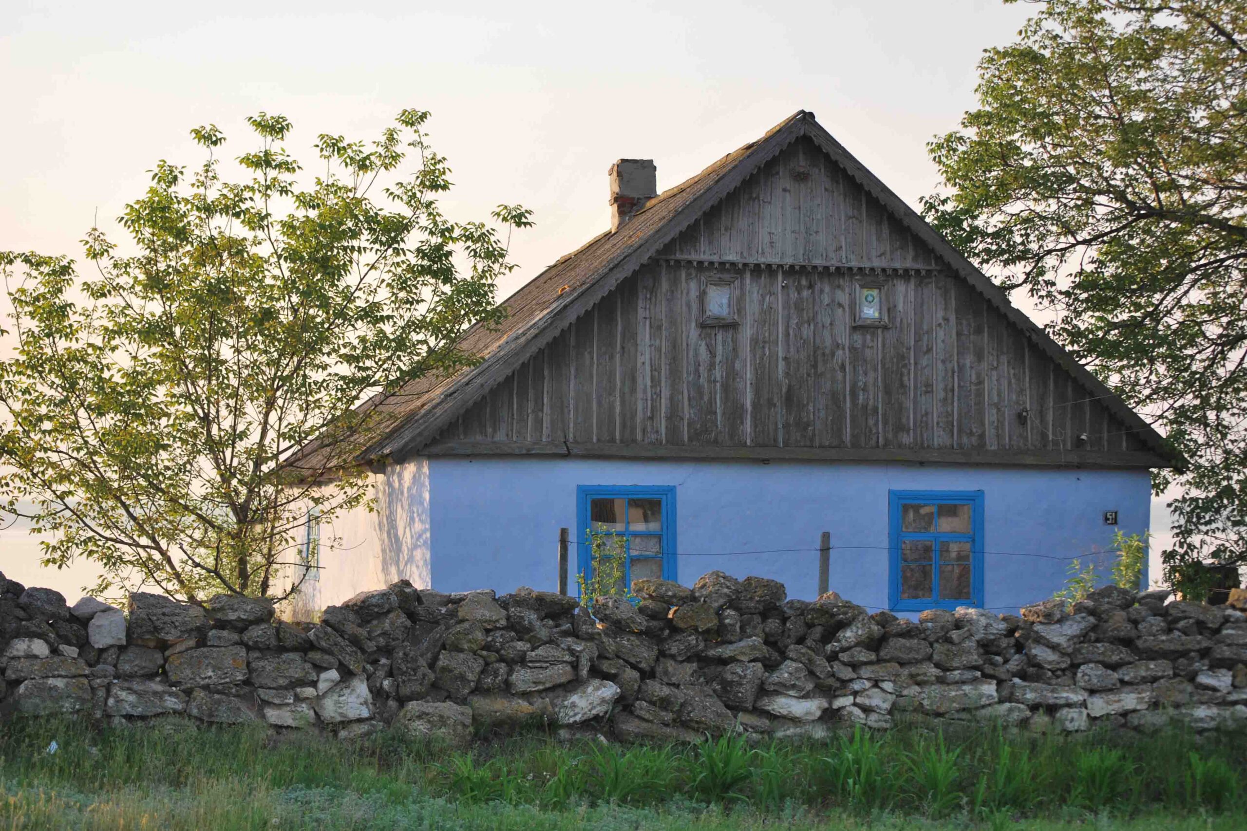 Ett litet blått hus med trätak står bakom en låg stenmur, med två fönster inramade i blått och ett träd som växer i närheten, under en delvis molnig himmel.