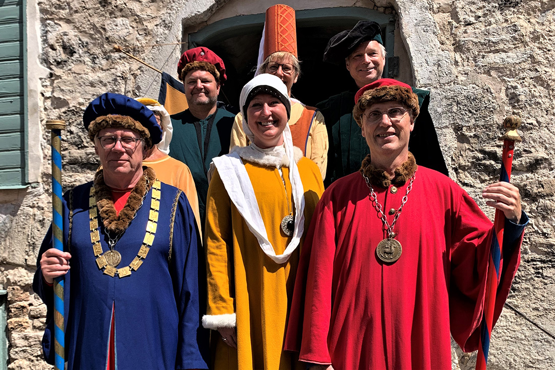 En grupp på sex vuxna i färgglada medeltida dräkter poserar utanför en stenbyggnad. De bär dräkter, hattar och medaljonger, står och ler i starkt solljus nära ett fönster och en välvd dörröppning.