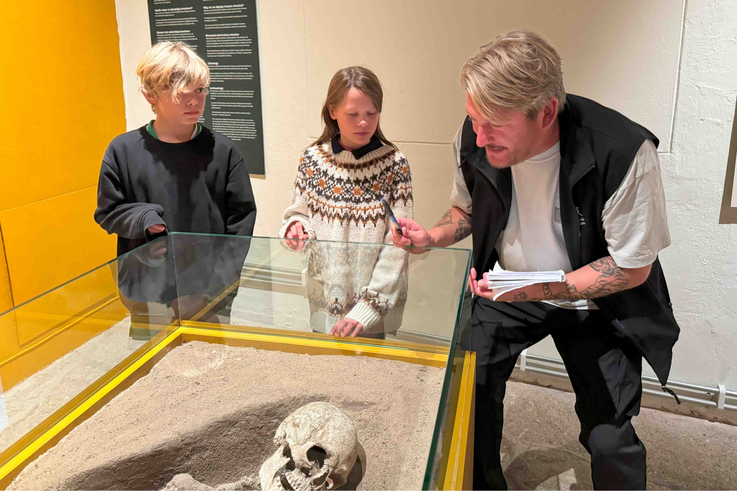 En man förklarar något för två barn medan han pekar på en mänsklig skalle som visas i en glasmonter på ett museum. Barnen lyssnar uppmärksamt, och informationsplaketter syns på väggen i bakgrunden.