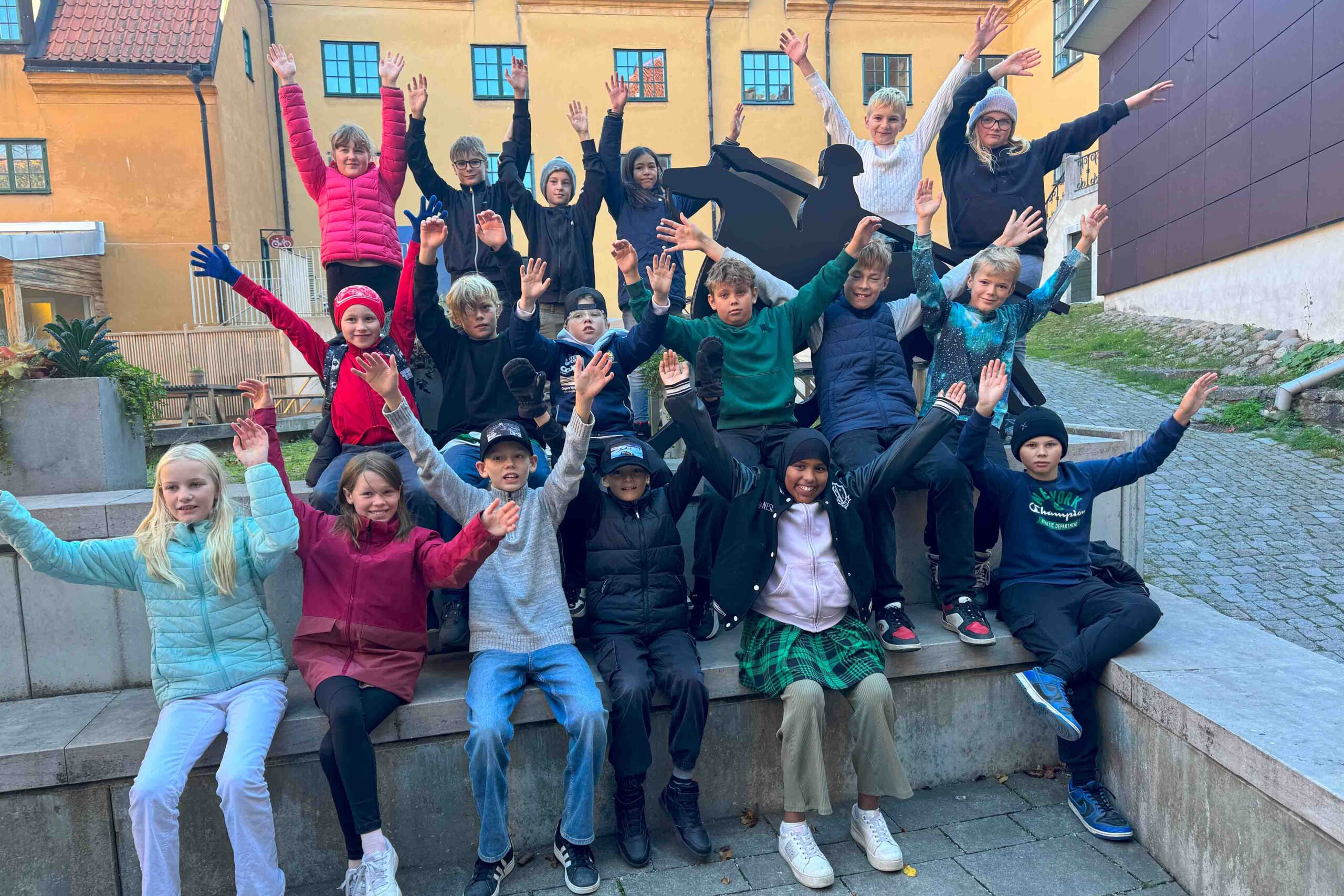 En grupp barn och tonåringar sitter och står på utomhustrappor, ler och sträcker armarna i luften. De bär jackor och mössor, vilket antyder svalt väder. Bakgrunden visar gula och bruna byggnader.
