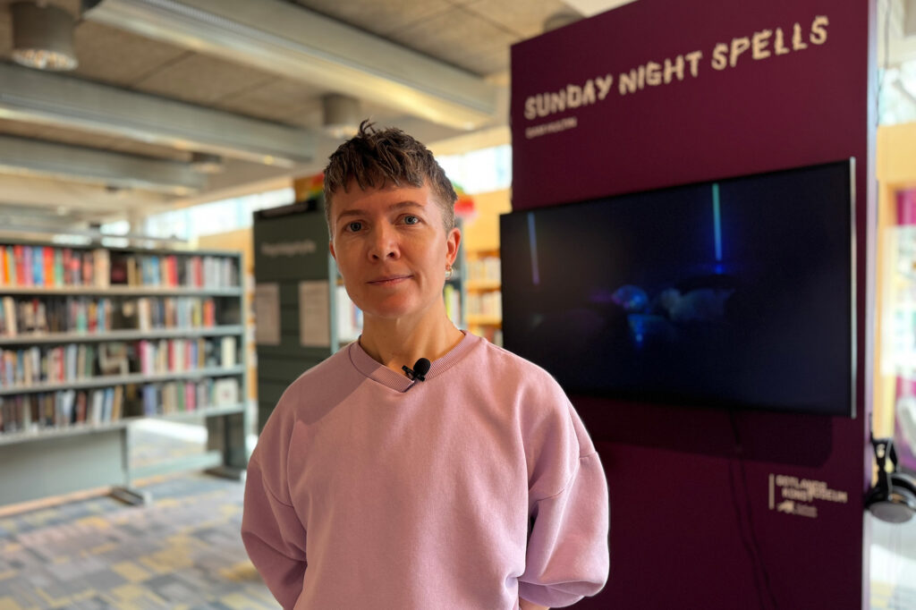 En person står i ett bibliotek med rosa tröja. Tittar in i kameran.