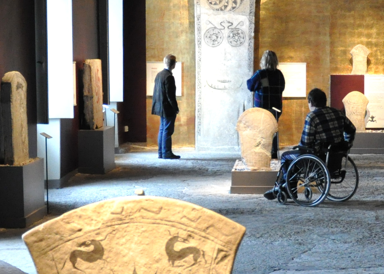 Tre personer, varav en i rullstol, tittar på antika stenartefakter och sniderier som visas i ett väl upplyst museumsgalleri med texturerade golv och väggar i varma toner.