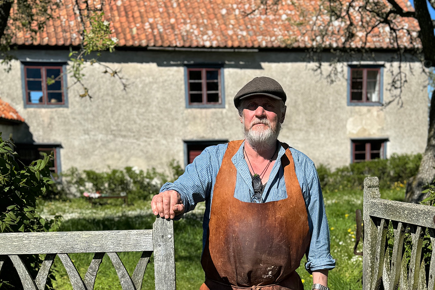 En man med en keps och förkläde i läder. Står vid en grind. Bakom sig har han ett hus ikalksten med tegeltak.