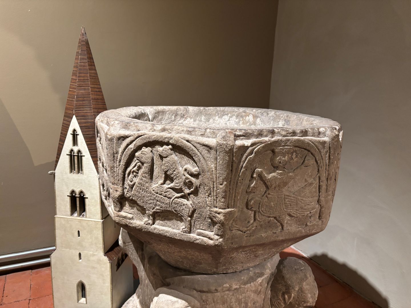 En dopfunt i kalksten. på ett museum. En modell av en kyrka bakom.