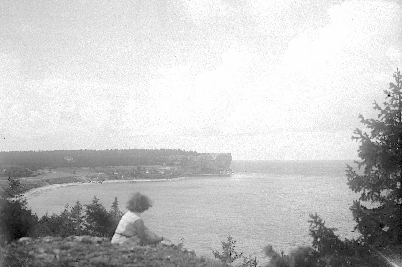 Svartvit äldre bild. Kvinna sitter på marken. Ser ut över havet och klintkust. Gotland.