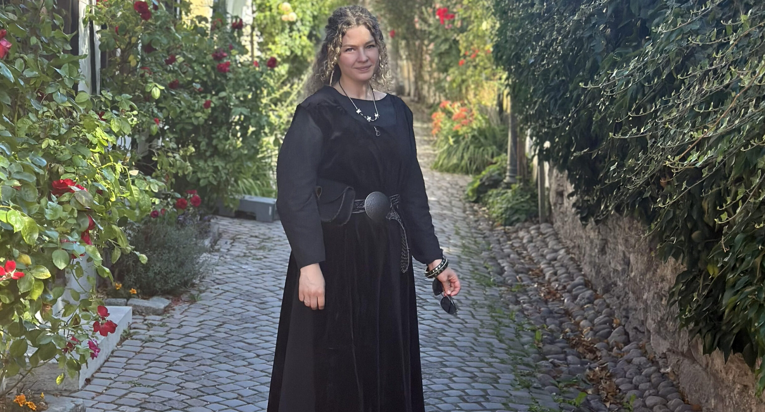 En kvinna med långt hår och medeltidsklänning. Står i en gränd med rosor. Visby. Sommar.