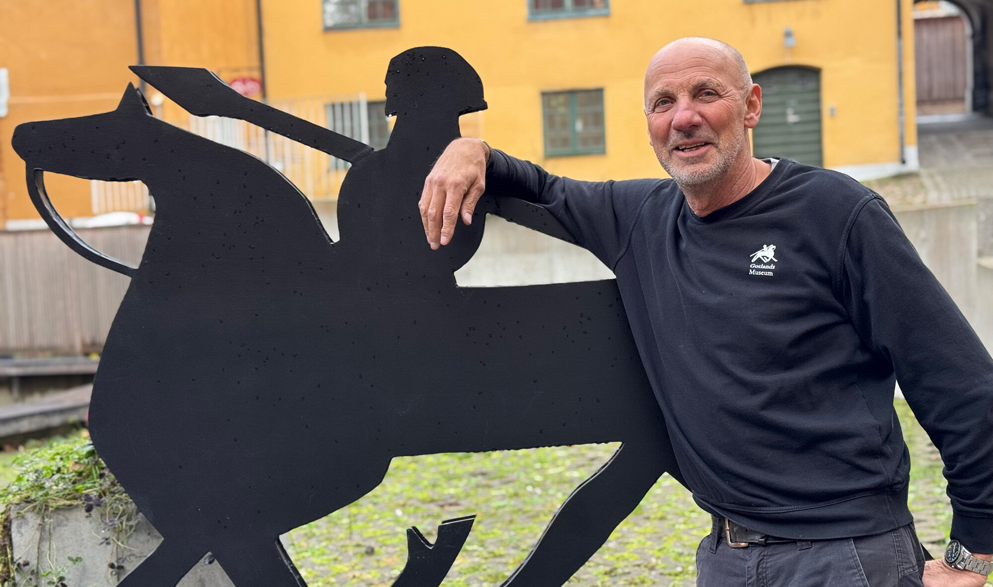 En man som står framför en vikingatida häst med ryttare och svärd. Han lutar sig mot hästen som är i svart smide.